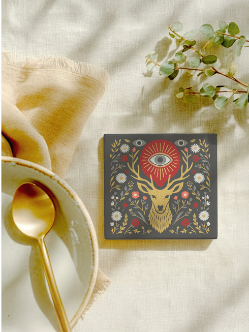Square ceramic coaster, 4x4 inches, with cork backing. Charcoal tile showcases a golden stag head with antlers, surrounded by florals and an all-seeing eye in red and gold. Smooth printed surface; bold folk-art style. Witchy occult deer coaster for dark academia décor, bar cart, or living room. Square ceramic drink coaster with cork base, stag and eye design.